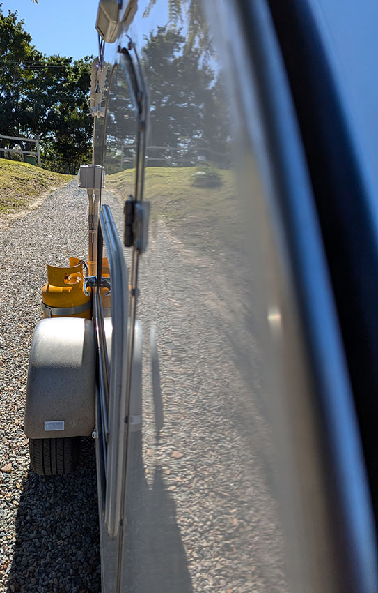 Side view of tear drop camper showing left wheel and entry door. A reflection on the side wall of trees.